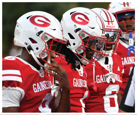 Three people wearing red Gainesville football jerseys and white helmets are standing closely together, appearing focused and ready for a game. Description generated by AI