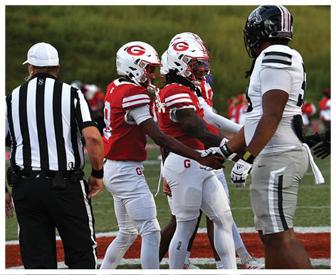 Two football players in red and white uniforms shake hands with a player in a white and black uniform while a referee stands nearby on the field. Description generated by AI