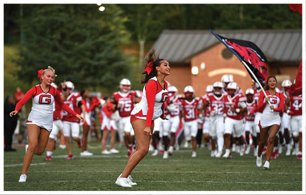 Three adults in cheerleading uniforms with red and white colors are on a football field with a group of football players in red and white uniforms running behind them, and one cheerleader is holding a large flag. Description generated by AI