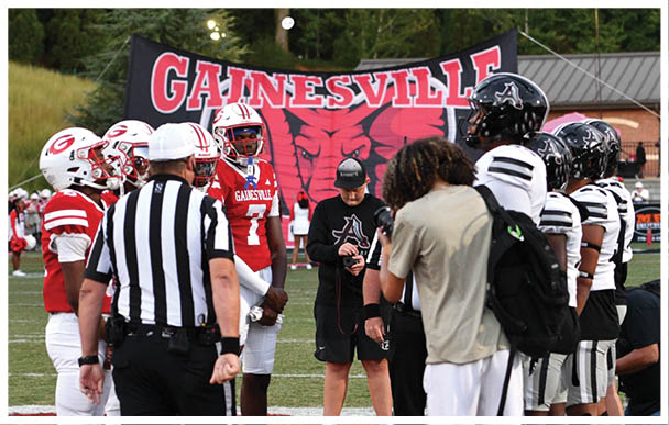 A group of football players and referees stand together on a field near a large banner that reads “Gainesville" with a red and black design in the background. Description generated by AI