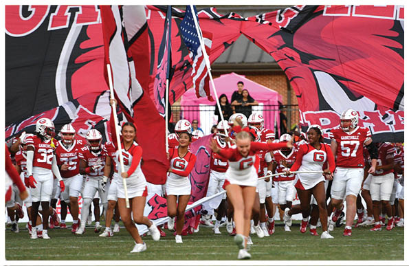 A group of people in sports uniforms and cheerleading outfits are running onto a field through a large inflatable tunnel with flags and a mascot logo. Description generated by AI
