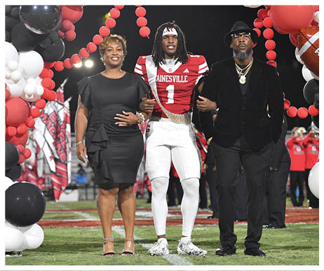 A person in a football uniform stands arm-in-arm with two people dressed in formal attire under a balloon arch decorated with football-themed balloons on a sports field at night. Description generated by AI
