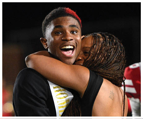 Two people dressed formally are embracing and smiling at an event with blurred people in red attire in the background. Description generated by AI