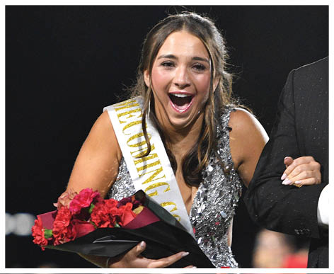 A person wearing a sparkling dress and a sash holding a bouquet of red roses looks excited while linking arms with another person dressed in a suit at an outdoor event with blurred people in the background. Description generated by AI