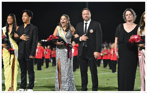Five adults dressed formally stand on a field with some wearing sashes and holding flowers while a group of people in red uniforms stand in the background. Description generated by AI