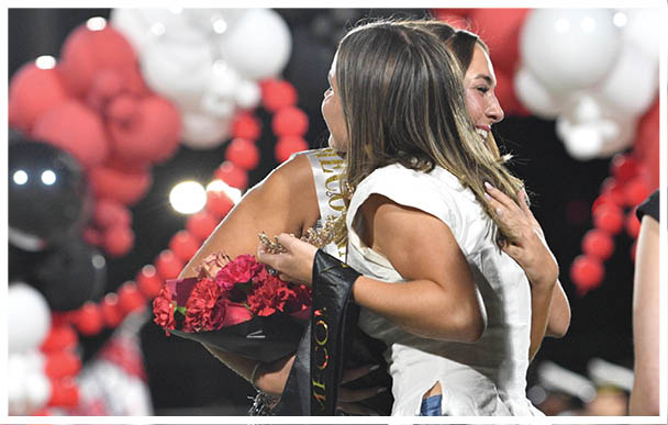 Two people are hugging warmly on a stage decorated with red, white, and black balloons, with one holding a bouquet of flowers and wearing a sash. Description generated by AI