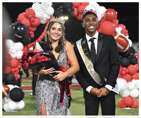 Two people dressed in formal attire wearing crowns and sashes stand on a football field with one holding a bouquet of flowers and smiling. Description generated by AI