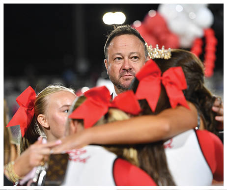 An adult with short hair and a beard looks thoughtfully while surrounded by people wearing large red bows and a crown. Description generated by AI