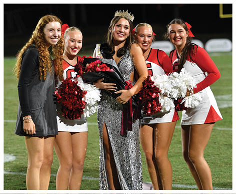 Five people stand together on a sports field with three wearing cheerleading uniforms holding pom-poms, one wearing a sparkly gown and crown holding flowers, and one wearing a casual dress, all smiling. Description generated by AI