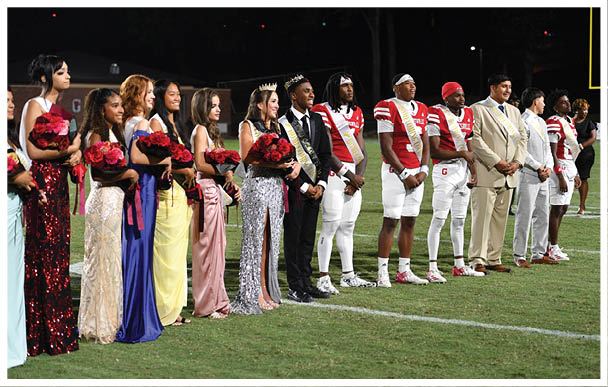 A group of people wearing crowns, sashes, formal attire, and football uniforms stand together on a field, smiling and posing for a photo. Description generated by AI