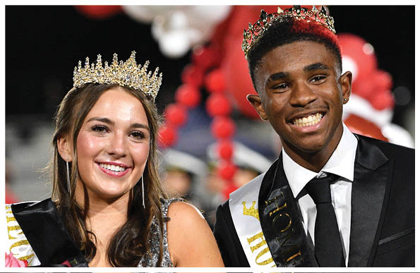 Two adults wearing crowns and sashes smile while standing together, with one holding a bouquet of red flowers, likely at a celebratory event such as a homecoming. Description generated by AI