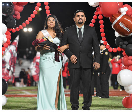 A person in a light blue formal dress holding a bouquet and wearing a sash stands arm-in-arm with a person in a black suit on a decorated field with red, black, and white balloons and blurred spectators in the background. Description generated by AI