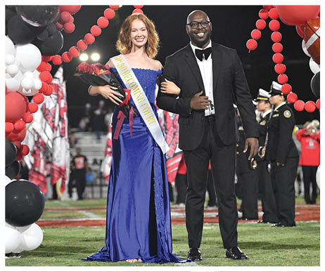 Two adults dressed formally stand arm in arm under red balloon decorations, with one wearing a sash that reads “Homecoming Court" and holding a bouquet of flowers. Description generated by AI