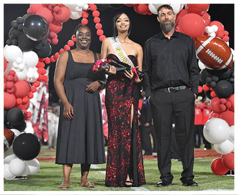 Three adults stand on a football field under a balloon arch decorated with football-themed balloons, with one person wearing a sash and holding flowers and a plaque. Description generated by AI