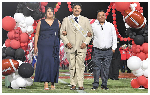Three adults dressed formally stand arm in arm under a balloon arch decorated with football-themed balloons on a sports field at night. Description generated by AI