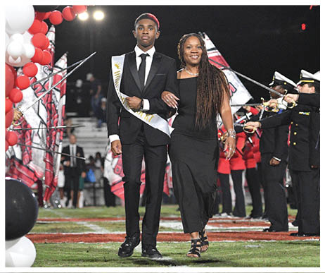 A person wearing a suit and sash walks arm-in-arm with another person in a long black dress on a decorated field with balloons and a marching band in the background. Description generated by AI