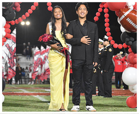 Two people dressed formally stand arm in arm on a football field under a decorative balloon arch with football-themed balloons, with one holding a bouquet of flowers and both smiling. Description generated by AI
