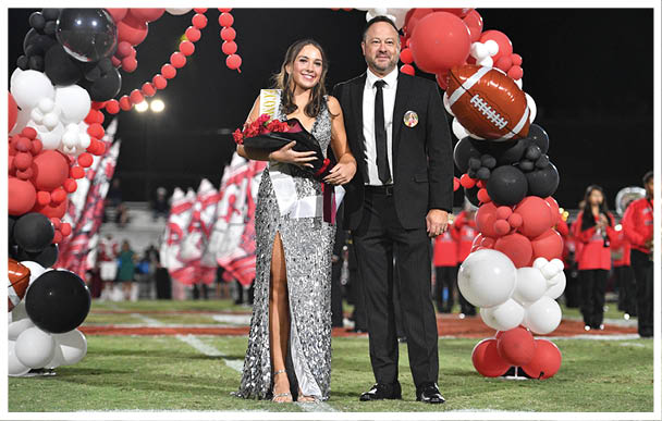 A person wearing a sparkly silver dress and sash holds a bouquet of flowers while standing next to a person in a black suit under a balloon arch decorated with red, black, and white balloons and a football-shaped balloon on a sports field. Description generated by AI