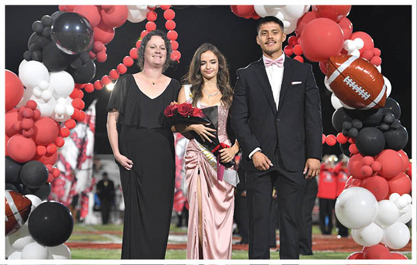 Three people dressed formally stand together with one holding a bouquet of roses and a sash, framed by red and black balloons including a football-shaped balloon. Description generated by AI