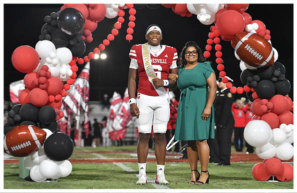 A person wearing a football uniform and a sash stands next to a person in a green dress under an arch of red, white, and black balloons at a nighttime event. Description generated by AI
