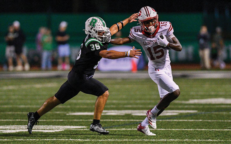Two people in football uniforms are actively engaged in a play on a football field, with one person reaching out to block or tackle the other who is holding the ball. Description generated by AI
