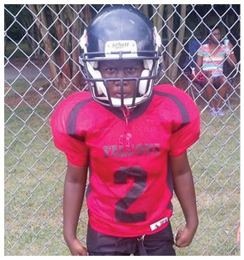 A child wearing a red football jersey with the number two and a black helmet stands in front of a chain-link fence with a person sitting in the background. Description generated by AI