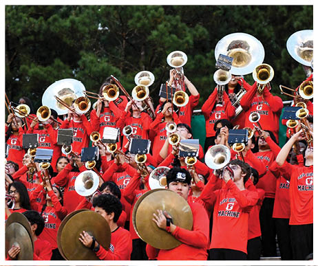 A large group of people wearing matching red shirts with “Big Machine" printed on them are playing brass instruments and cymbals outdoors in a band setting. Description generated by AI