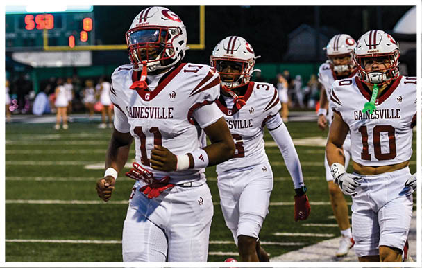 Three people in white football uniforms with “Gainesville" on the jerseys are running on a football field during a game. Description generated by AI