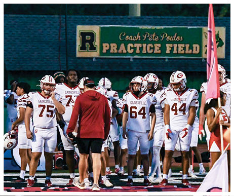Several people in football uniforms stand on a practice field under a sign that reads Coach Pete Poulos Practice Field while a person in cheerleading attire holds a flag in the foreground. Description generated by AI