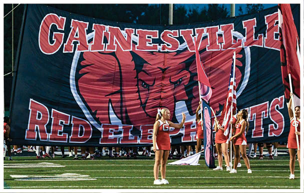 People in red cheerleading uniforms hold flags in front of a large banner with an elephant and the words “Gainesville Red Elephants" at a sports event. Description generated by AI