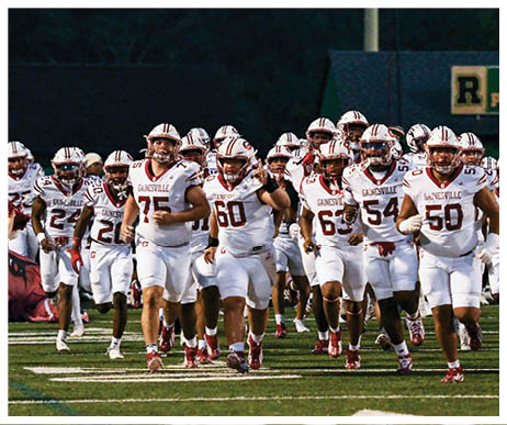A group of football players in white uniforms with “Gainesville" on their jerseys are running onto a field accompanied by a cheerleader in a red outfit. Description generated by AI
