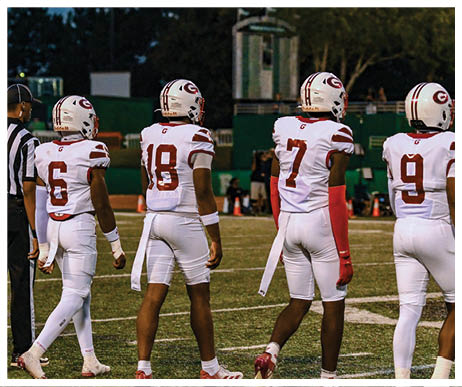 Three people wearing white football uniforms with helmets stand on a field during a game or practice. Description generated by AI