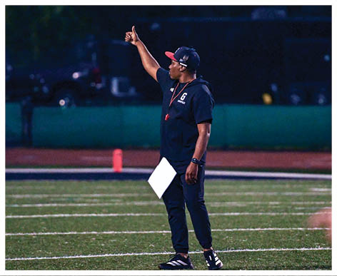 A person wearing a cap, dark athletic clothing, and sneakers stands on a sports field holding a clipboard and giving a thumbs-up gesture. Description generated by AI