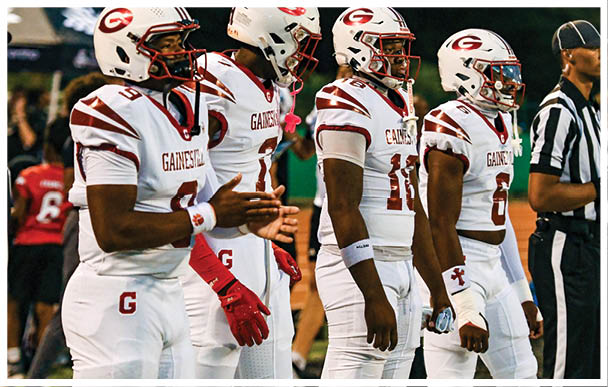 Four football players in white uniforms with red accents and helmets stand side by side on a field with a referee nearby. Description generated by AI
