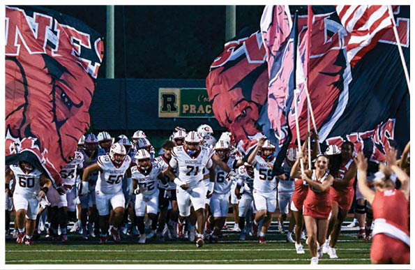 A group of athletes in white football uniforms run onto a field behind large red and black flags while people in red outfits cheer and wave flags. Description generated by AI