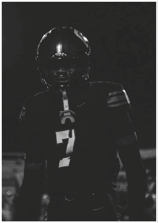 A person wearing a football uniform with the number seven stands in a dark setting, illuminated slightly from the front. Description generated by AI
