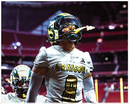 A person wearing a football helmet and a white jersey with the word Grayson stands on a field inside a stadium. Description generated by AI