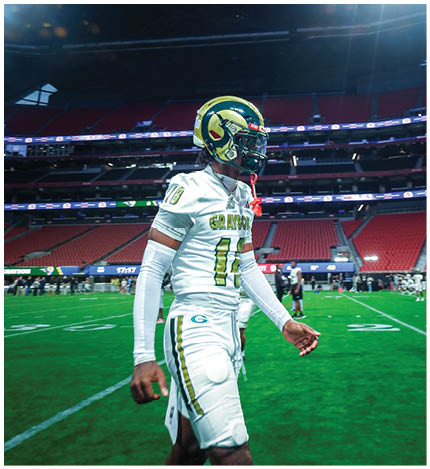 A person in a white football uniform with the number 10 and a gold helmet stands on a football field inside a stadium with a scoreboard displaying “Classic" above. Description generated by AI