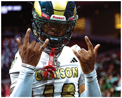 A person wearing a football helmet and white jersey with “GRAYSON" written on it is making hand gestures with both hands. Description generated by AI