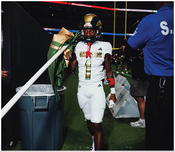 A person in a white football uniform holding a flag stands on a field near a trash can and other people. Description generated by AI