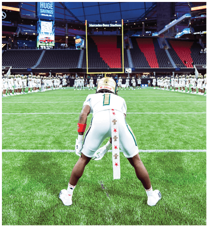 A person in a white football uniform is positioned on a green field facing a goalpost inside a large stadium with empty seats and other players lined up in the background. Description generated by AI
