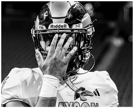 A person wearing a football helmet and uniform is holding their hand up to their face in a black and white photo. Description generated by AI