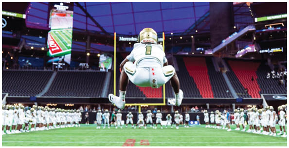A person wearing a football uniform with the number one is jumping in the air inside a stadium with a partially closed roof and empty seats. Description generated by AI