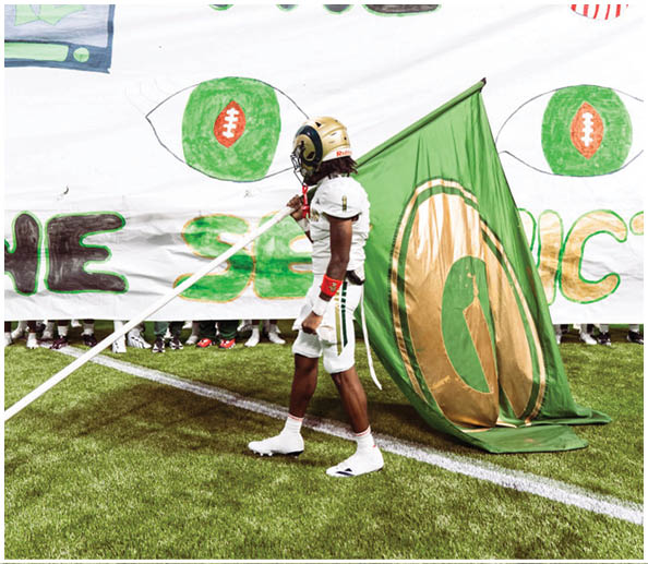 A person wearing a helmet and holding a green flag stands in front of a banner with large green and black letters and football-shaped designs. Description generated by AI