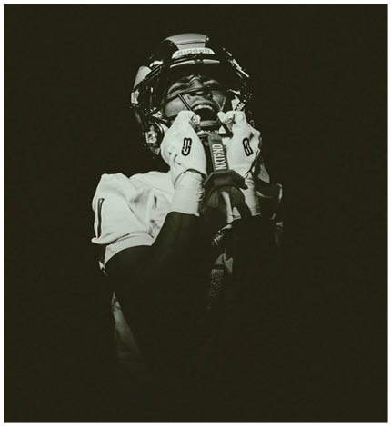 A person wearing a football helmet and gloves is captured in a dramatic black and white photo, appearing to shout or express intense emotion. Description generated by AI