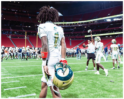 A person with dreadlocks wearing a white sports uniform with the number 1 stands on a field inside a large stadium with bright lights and other people in sports attire in the background. Description generated by AI
