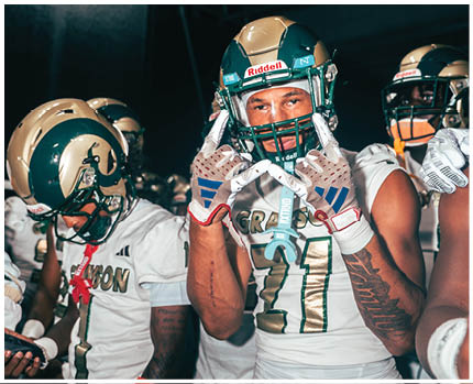 A group of people wearing football helmets and uniforms are gathered closely, with one person in the center making a hand gesture. Description generated by AI