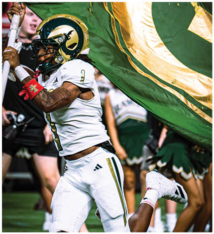 A person in a white football uniform and helmet is running while holding a large green and gold flag with a letter G, with cheerleaders visible in the background. Description generated by AI