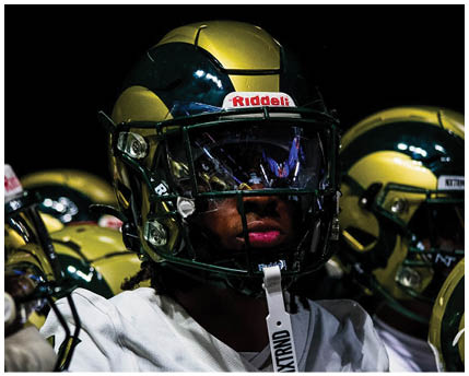People wearing football helmets and uniforms are closely gathered, with one person in the foreground looking focused. Description generated by AI