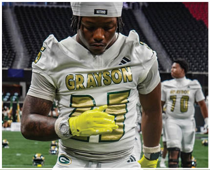 A person wearing a white football uniform with “GRAYSON" and the number 25 on it is looking down while wearing a white cap and bright yellow gloves, with another person in a similar uniform in the background. Description generated by AI